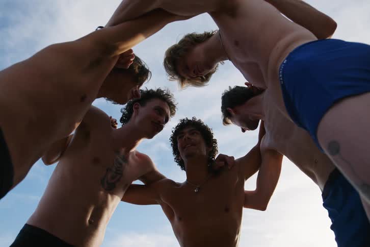 Male friends talk in a huddle on the beach then walk to the water on a cloudy summer day