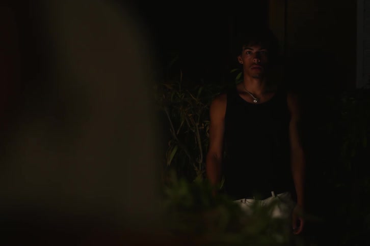 A man wearing a tank top and necklace appears and starts looking straight ahead at something that is out of focus and then leaves and it is dark