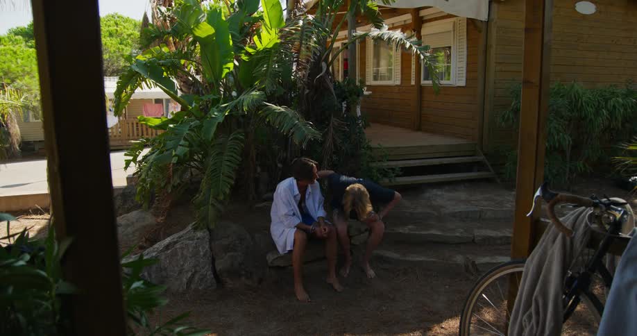 A man wearing headphones and a red shirt walks to a wooden porch where some friends are sitting and talking