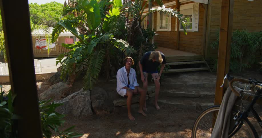 A boy in a red shirt comes out of a house and goes to the porch where there are some friends sitting and talking and then two other friends walk by talking in the sandy yard and one of them sits on a rock