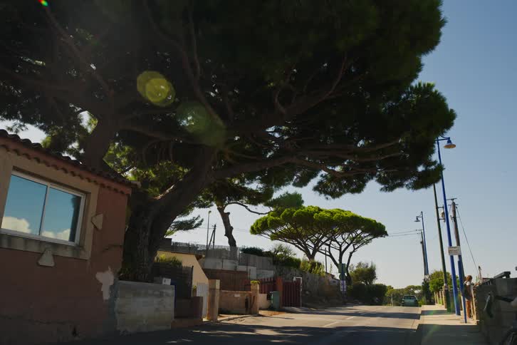 Two cars driving down a street with houses and trees on both sides and people walking on the sidewalks