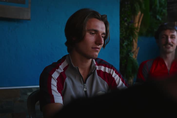 A young man is drinking from a glass and then has a sad face and behind him there is a man talking