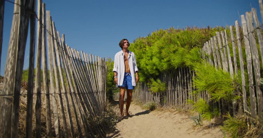 A teenage boy walks down a path at a beach and then stops and thinks