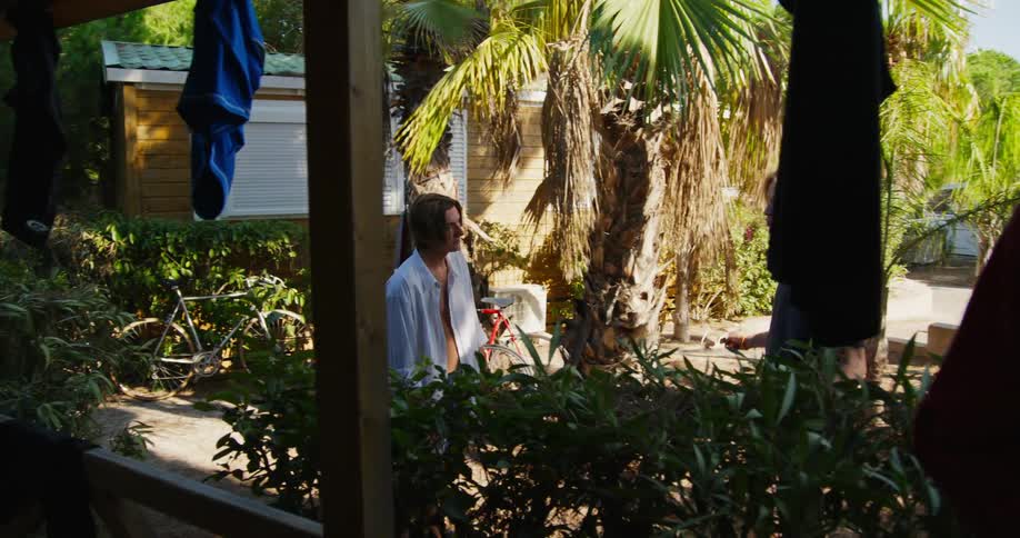 Young friends talking and drinking at a table on the porch of a resort house and two other friends arriving on bicycles and sitting on a rock and talking to each other