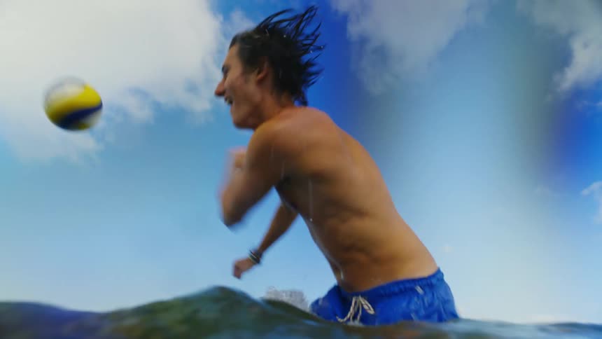 A young man hits back a volleyball while standing in the water at a beach