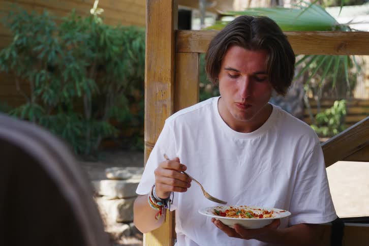 Male in a white tshirt sitting outdoors in a tropical location eating spaghetti