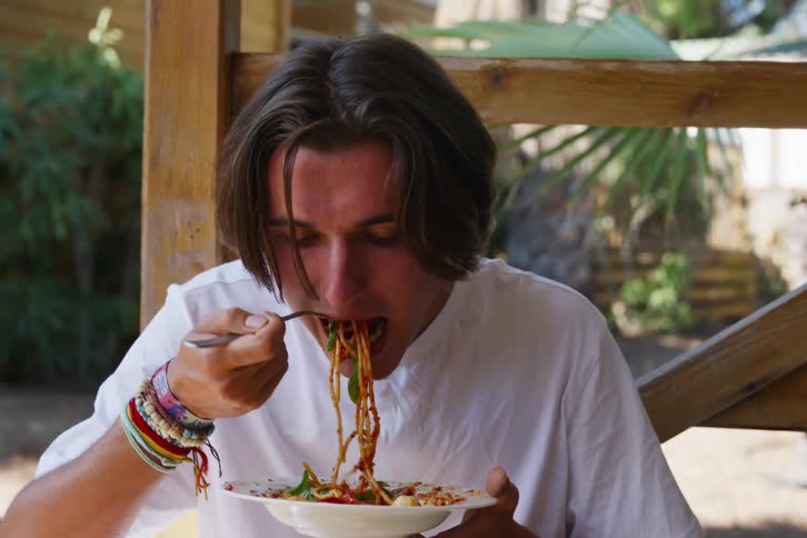 A young man sits on a patio while eating spaghetti from a bowl