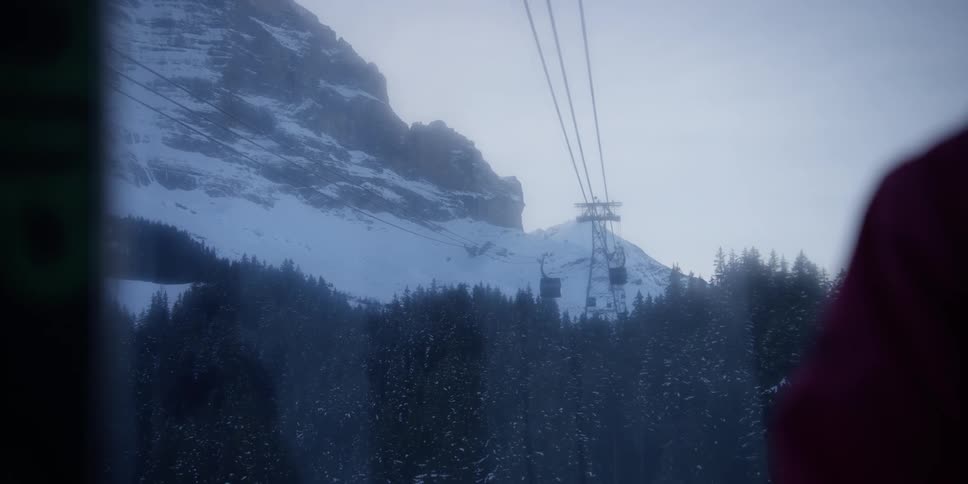 A cable car passing over trees at the base of a rocky mountain covered in snow