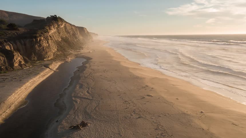An aerial shot of cliffs beside the ocean on a sandy beach at dawn