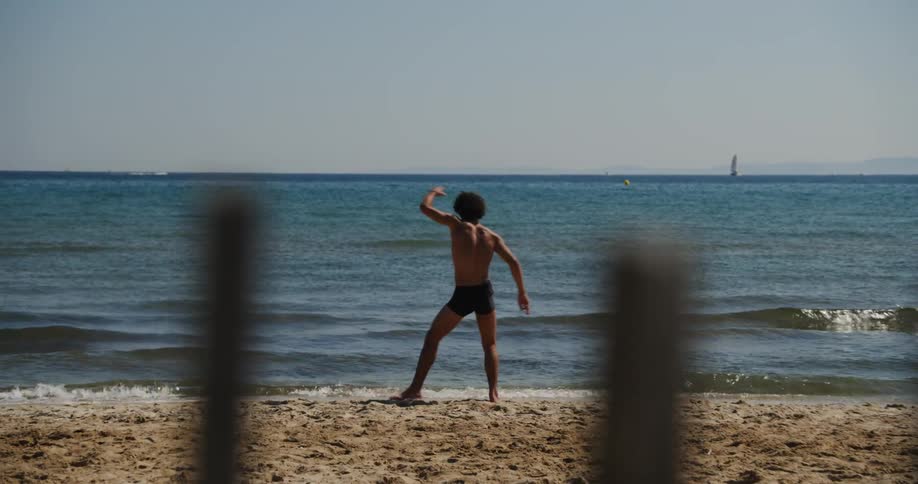 A young man is dancing in front of the beach and he is wearing only underwear and in front there are some wooden pillars