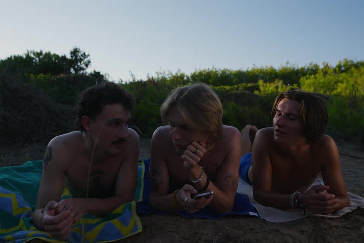 Man in swimming trunks on the sand talking on his cell phone and playing with an object with his colleague while his other friends are lying on the sand with a towel