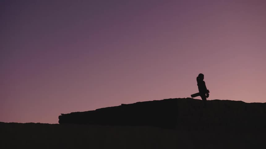 Silhouette of a girl on a hilltop picking up a board and walking in front of a purple sky at twilight