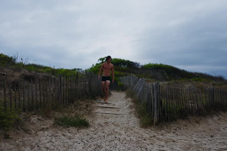 Two young men men walking along a sand walkway leading to the beach in just swim trunks on a cloudy day