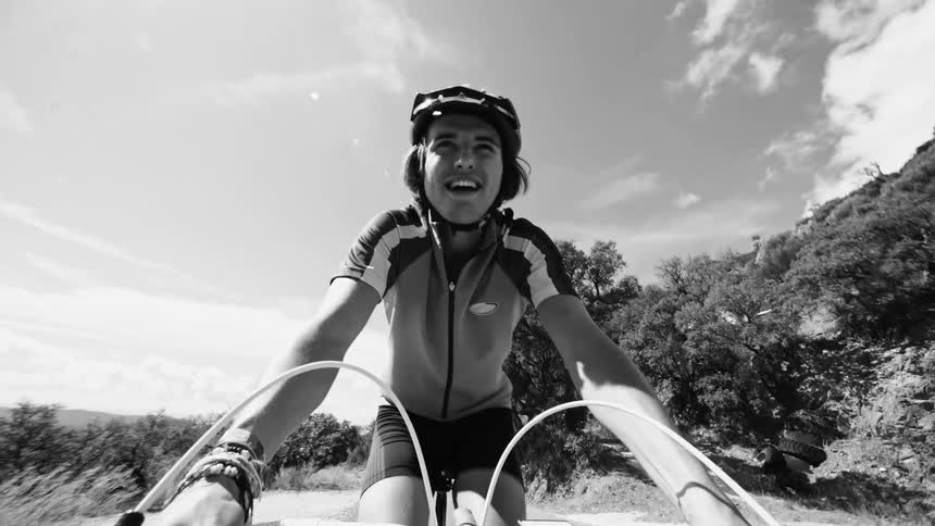 A man is riding a bicycle in a region of nature and is smiling and the tones are monochrome