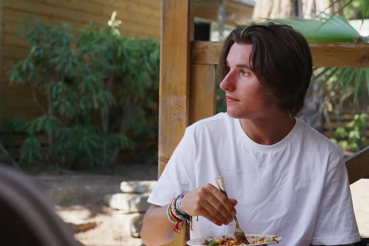 Portrait of a young man eating pasta while talking to his colleague and then other men walk by him and he stares
