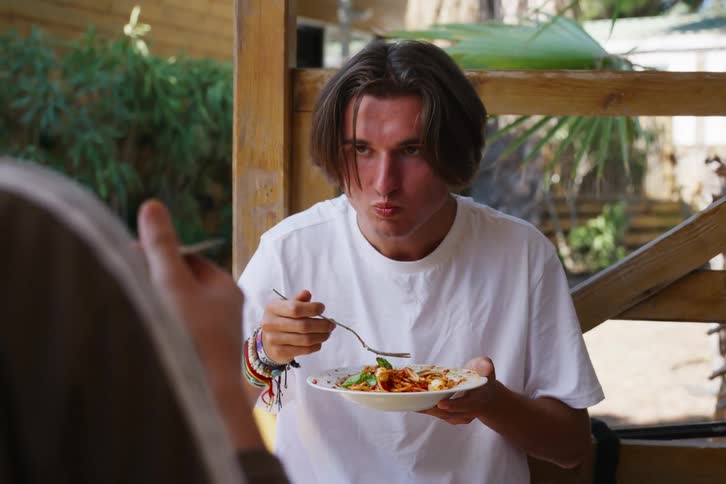 Male in white tshirt sitting outdoors on the lanai eating spaghetti with a friend