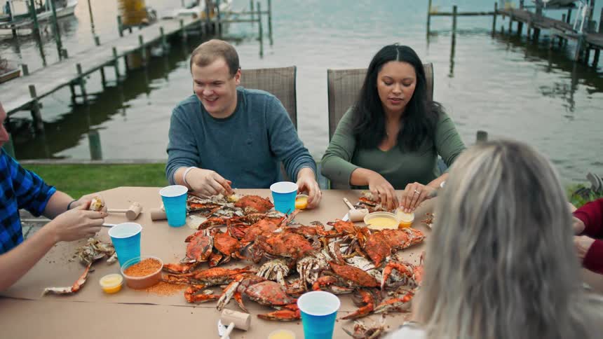 Several people are gathered at a table and talking while peeling and eating crabs