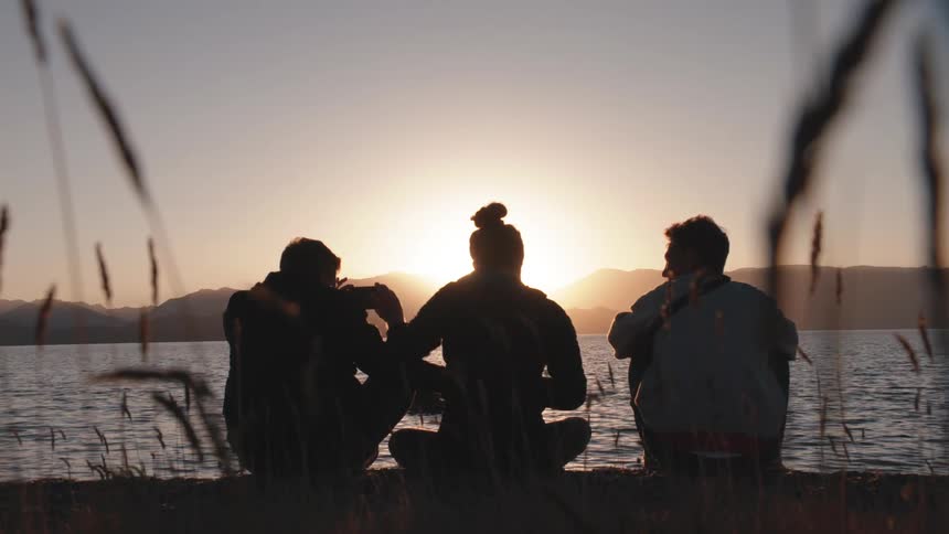 A woman and two guys sit by a lake and laugh and have some fun
