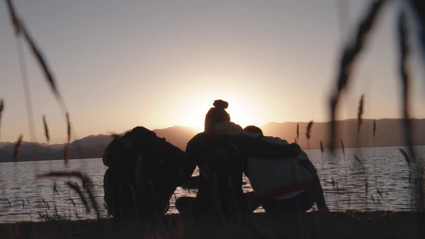 Three friends hug each other while sitting next to a lake at sunset