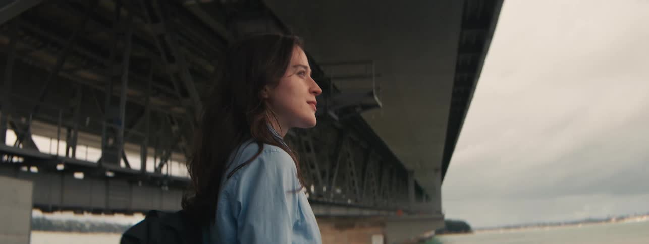 A woman with a black backpack is happily walking under a bridge over a bay
