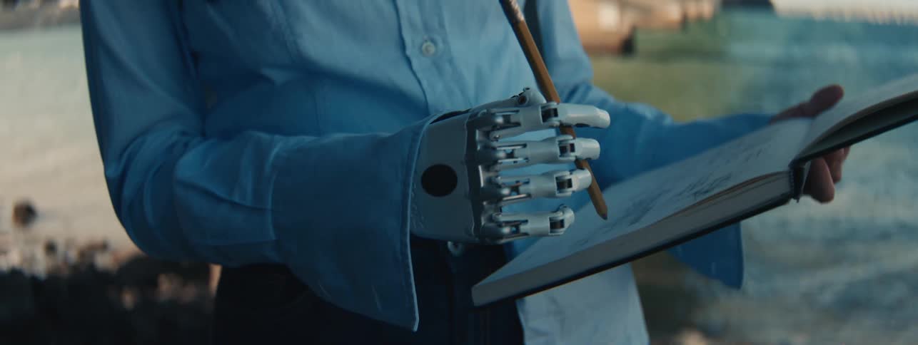 Close up of a woman with a prosthetic robotic arm writing on a notepad near a lake
