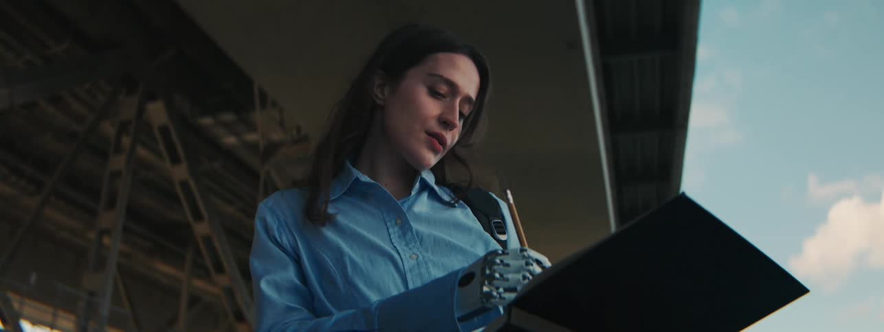 A woman standing outdoors uses a mechanical hand to write in pencil on a notepad