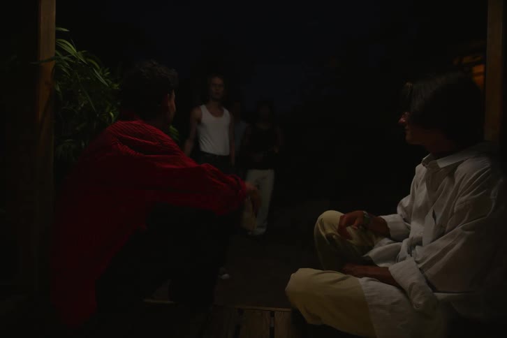 Two young people sitting outside the house one of them reading a book and the other playing the guitar and then a group of friends arrive with drinks and they enter the house