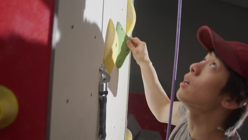 A young man is climbing a wall using a safety rope and hanging from colorful climbing holds on the wall