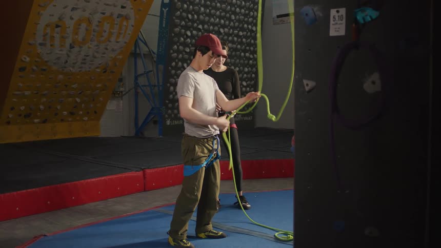 A man tying a safety rope in a climbing gym as a woman observes