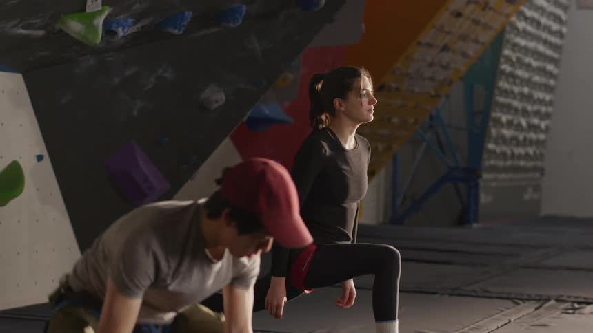 A man and a woman in a gym full of wall climbing gear doing stretches