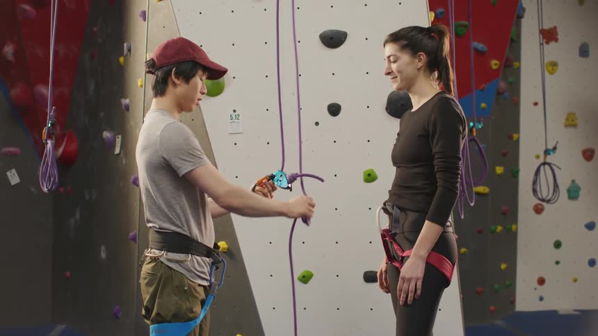 An asian man shows a young woman how to tie a rope in a climbing gym