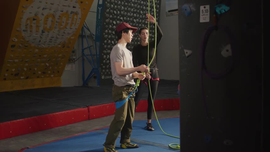 A man climbs the wall at an indoor climbing center as a woman stands below holding the safety rope