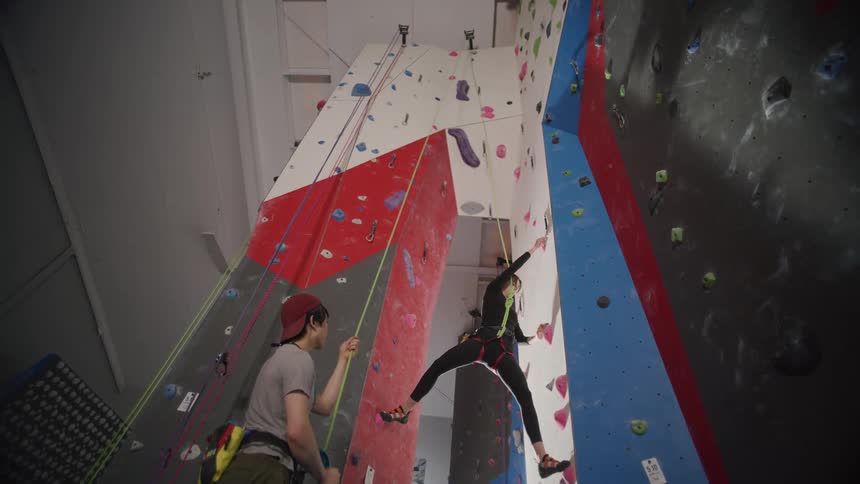 A woman practicing wall climbing in a gym while a man holds her security rope