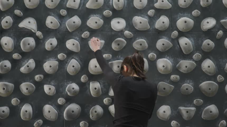 A tall woman begins to ascend an indoor climbing kilter board with a variety of holds