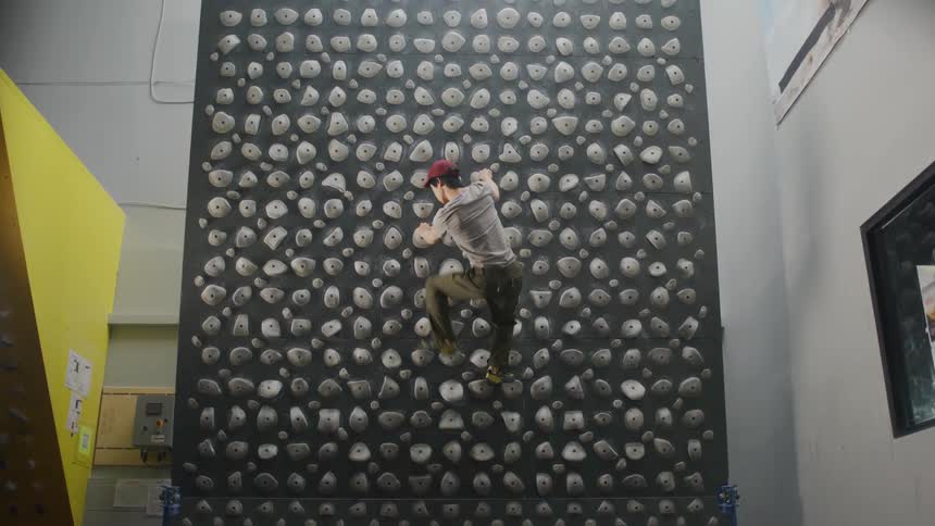 A young man in a red cap trains on a kilter board climbing wall with a variety of hold types