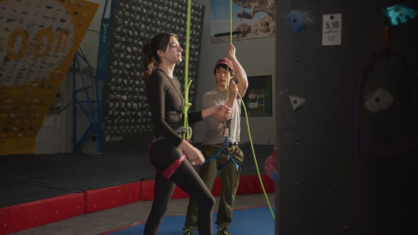 Man and woman side by side both with ropes and a safety belt and then the woman begins to climb training on a fake indoor climbing wall