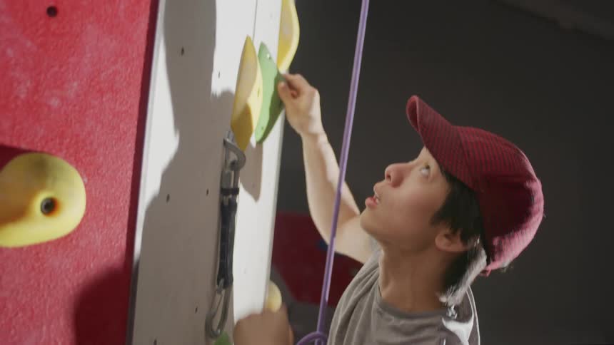 A young man in a red cap is climbing a wall of climbing holds using a purple rope