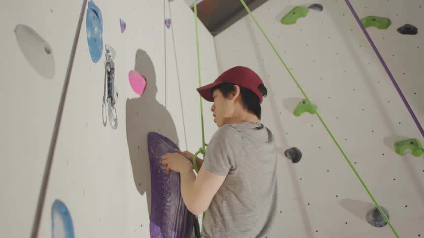 A young man in a red cap is climbing a wall with colorful climbing holds and using a green protective rope