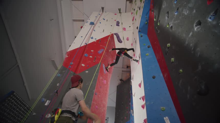 Woman climbing a climber wall with the help of a man holding a safety harness