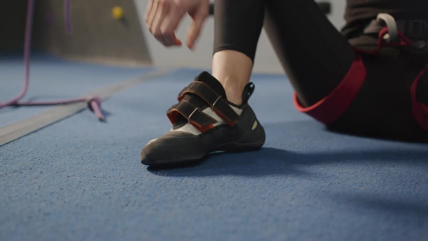 A person is sitting on a blue floor tying the straps of their climbing shoes