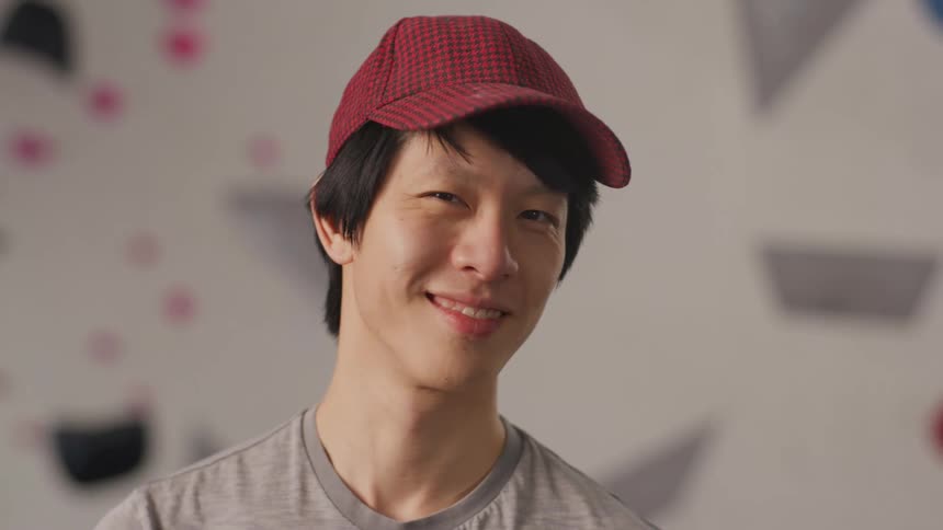A young asian man wearing a red cap looking to the side then looks straight ahead and starts to smile