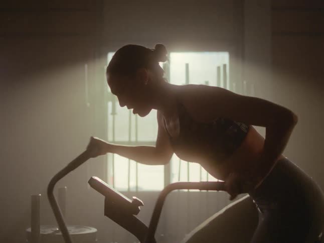 A female athlete woman works out on a stationary bike in a gym with epic lighting behind her