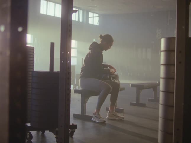 A tall woman walks into a gym and sits on a bench as she sets down her bag