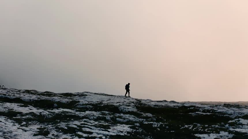 The silhouette of a man wearing a backpack hiking along a snow dusted black rock ridge