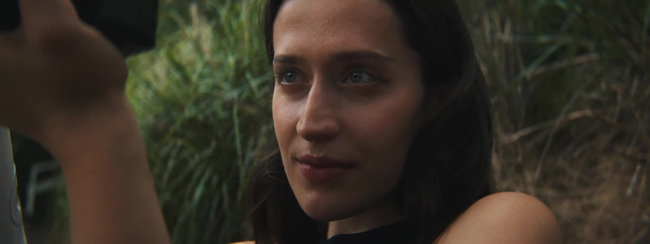 A woman holding a camera to her face outdoors with vegetation visible in the background
