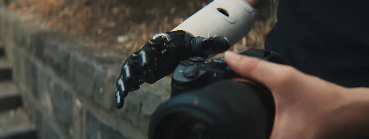 A human hand holding a camera and on the other side a prosthesis closing the hand to hold it too
