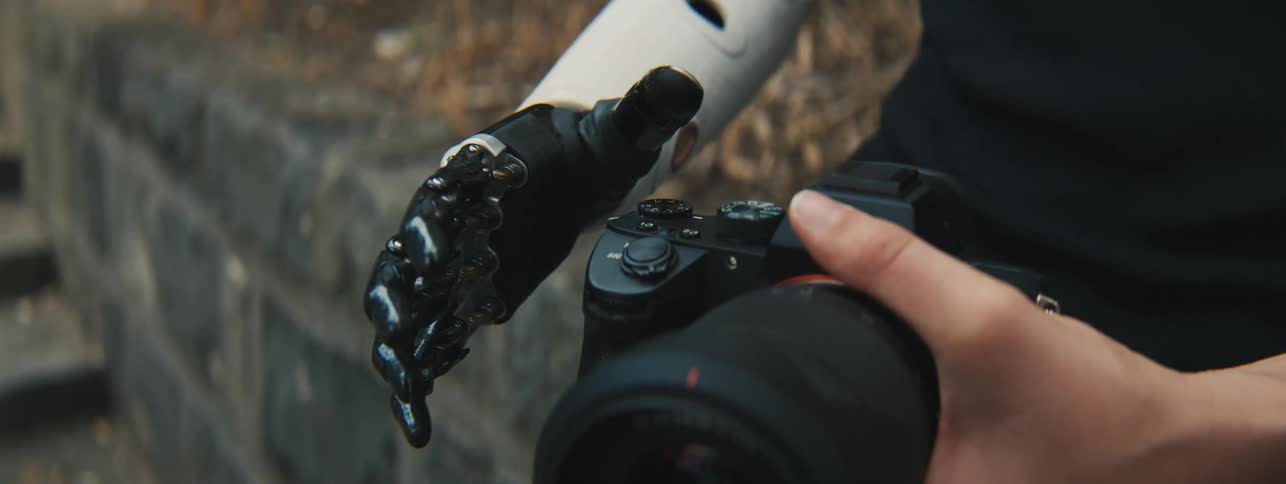 A prosthetic hand opening to hold a digital camera that is in a persons human hand