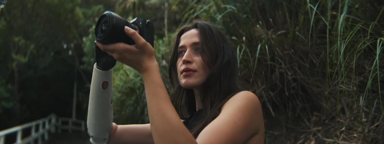 Lady holds a camera up while standing on a stair near the bush at midday