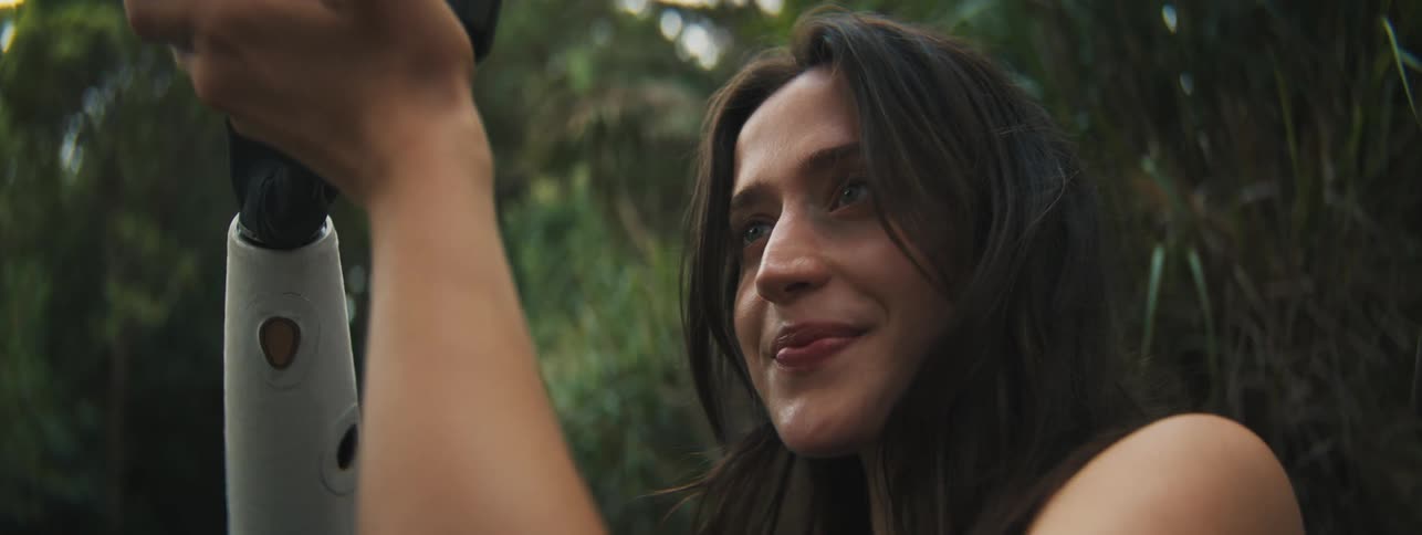 Young woman smiling slightly while holding a professional camera in an environment with outdoor vegetation