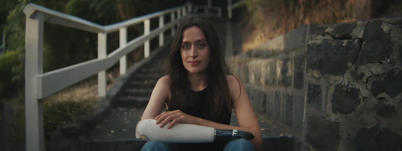 A young woman with a prosthetic arm is sitting on stone stairs outside